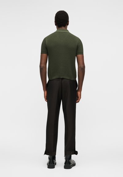 Άνδρας που στέκεται με την πλάτη προς τα έξω, φοράει ένα κοντομάνικο πλεκτό πουκάμισο σε ελαιοπράσινο χρώμα, μαύρο παντελόνι με ιμάντες στριφώματος και μαύρα δερμάτινα παπούτσια.