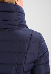 Mörkblå puffjacka med hög krage, med quiltad stoppning. Texturerad yta med horisontell sömnad och en liten logotypaccent.