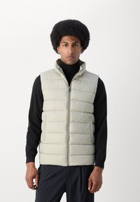 Man wearing beige quilted sleeveless vest over black turtleneck and pinstriped pants, standing against plain white background.