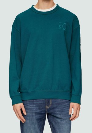 Homme portant un sweat-shirt à col rond turquoise avec un petit logo "s.O" sur la poitrine et un jean bleu.