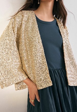 Kobieta w czarnej sukience z luźną, złotą cekinową kurtką i perłowymi kolczykami wiszącymi, ręka spoczywa na spódnicy.