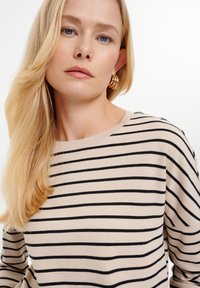 Beżowy sweter w paski z czarnymi poziomymi pasami. Miękka struktura, okrągły dekolt i delikatne szwy na ramionach. Modelka z blond włosami i złotymi kolczykami w kształcie kół.