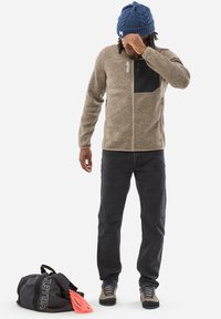 Veste en polaire marron avec poche poitrine noire, jeans noirs, chaussures grises avec lacets bleus, et un bonnet en tricot bleu, accompagnés d'un sac de sport noir.