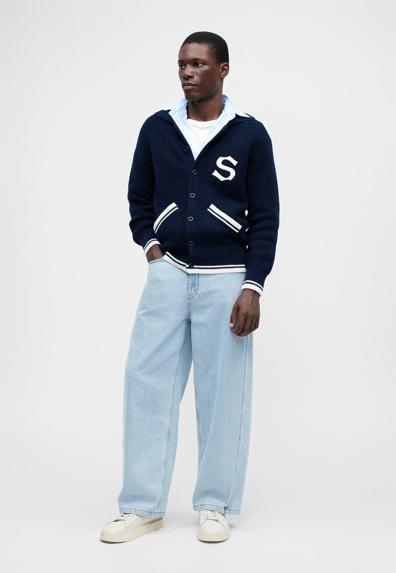 Marine gebreide cardigan met witte streepaccenten, voorzien van een logo en knopen, gedragen over een lichtblauw overhemd met kraag en losse lichtblauwe jeans.