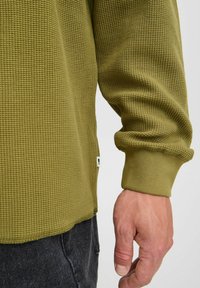Cămașă lungă cu mânecă, texturată în verde măsliniu, cu un model cu nervuri și mâneci cu manșete, având o etichetă mică pe tivul lateral.
