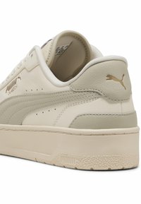 Tenis Puma Cali Match, de culoare bej și alb murdar, cu talpă joasă, parte superioară din piele și logo-ul Puma aurit pe toc și pe lateral, cu talpă texturată vizibilă.