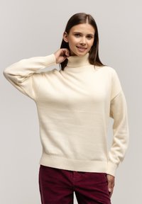 Sweter w kolorze kremowym z golfem, o luźnym kroju, z prążkowanymi mankietami i dołem, zestawiony z burgundowymi spodniami z kordurowej tkaniny. Gładka tekstura.