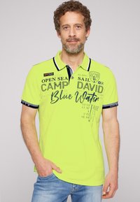 Heldergele polo shirt met marineblauwe accenten, voorzien van tekst en motieven. Korte mouwen met een kraag en een casual pasvorm. G gedragen met blauwe jeans.