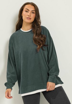 Femme aux longs cheveux bruns ondulés portant un sweat-shirt vert foncé surdimensionné par-dessus une chemise blanche et des leggings foncés, se tenant devant un mur clair et uni.
