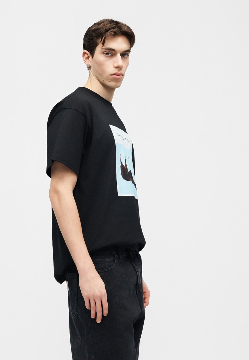 Junger Mann trägt ein schwarzes T-Shirt mit Grafik und schwarze Jeans, steht seitlich vor einem schlichten, hellen Hintergrund.