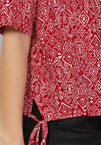 Rood en wit gebloemd shirt met korte mouwen, voorzien van geometrische ontwerpen en een strikdetail aan de onderkant. Gladde stoftextuur.
