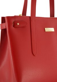 Rote, strukturierte Leder-Tote mit zwei Schulterriemen, goldener Hardware und einer kleinen goldenen Markenplatte. Verfügt über einen Reißverschluss und ein minimales Design.