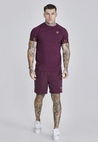 Bordowy t-shirt sportowy z krótkim rękawem i logo, zestawiony z bordowymi szortami. Widoczne tatuaże i białe trampki.