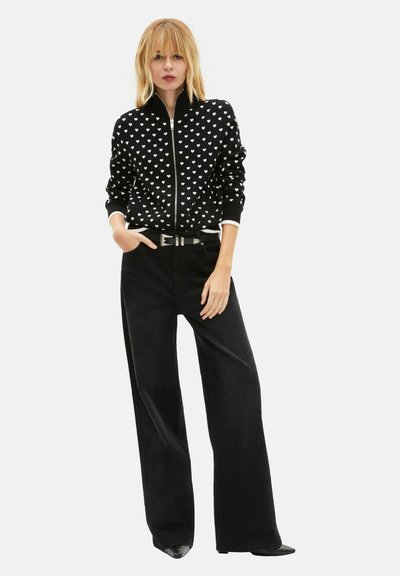 Pull noir zippé avec motifs de cœurs blancs, associé à un pantalon noir à jambes larges. Tissu texturé, silhouette ajustée et poignets blancs contrastants.