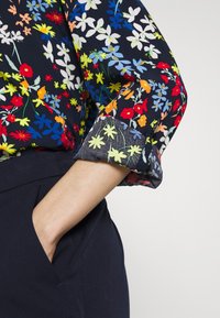 Kwiatowa bluzka na ciemnoniebieskim tle, ozdobiona wielokolorowymi wzorami kwiatów. Rękawy są mankietowe, odkrywając przezroczystą teksturę.