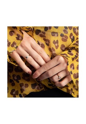 Two rings shown on hands: one gold with a gemstone accent, the other a delicate band. Background features a yellow and brown leopard print blouse.