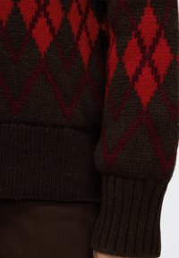 Gros plan d'un poignet de pull en tricot avec un motif losange rouge et brun foncé et un ourlet en côtes sur le bras d'une personne.