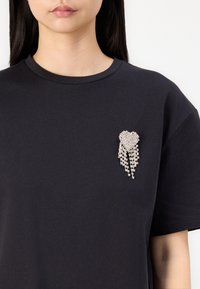 Femme portant un t-shirt noir uni avec une broche en forme de cœur argentée ornée de strass et des chaînes pendantes sur le côté gauche de la poitrine.