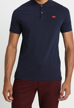 Mężczyzna ubrany w granatową koszulkę polo z krótkim rękawem z czerwoną wyszywaną naszywką i ciemnoczerwone spodnie, stojący z rozluźnionymi ramionami.