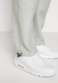 Ljust grå sweatpants med sid sömmar och svarta dragskoaccenter, kombinerade med vita Nike Air Max sneakers med en texturerad yta.