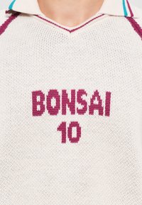 Krémszínű kötött póló ing V nyakkal, barna "BONSAI" és "10" felirattal. Törökzöld csíkok díszítik a gallért.