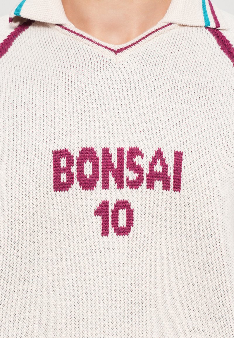 Polo en tricot crème avec un col en V, comportant le texte maroon "BONSAI" et "10". Agrémenté de rayures teal sur le col.