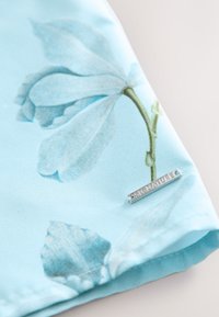 Țesătură albastru deschis cu un model floral cu petale moi și un tulpină verde. O etichetă argintie cu inscripția "SIGNATURE" este fixată pe margine.