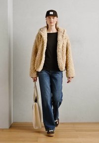 Kvinne som går i en beige, fluffy jakke, svart skjorte, blå jeans, mørk caps og sneakers, og bærer en beige tote-veske innendørs.
