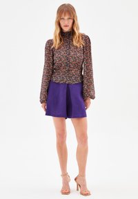 Blomstret bluse med lange ærmer og flæser ved håndleddene, parret med højtaljede lilla shorts. Modellen har nude stroppede hæle på.