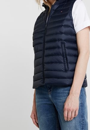 Femme portant un gilet sans manches matelassé bleu marine sur une chemise blanche à manches courtes et un jean bleu, debout avec les bras détendus.