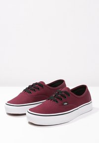 Burgunderfarbene Leinensneaker mit schwarzen Schnürsenkeln, weißen Gummisohlen und einem schwarzen Streifen um die Sohle, ausgestattet mit einem kleinen Logotag an der Seite.