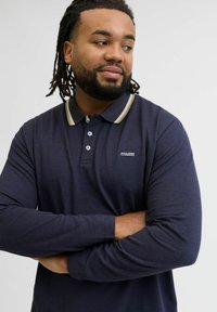 Vīrietis ar bārdu un dredsām, ģērbies tumši zilas krāsas garām piedurknēm polo krekla ar bēša krāsas apkakli, ar rokām saliktām krustām un skatoties uz sāniem.