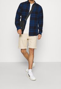 Camicia di flanella a quadri blu e neri, t-shirt bianca, pantaloncini beige, scarpe da ginnastica bianche con dettagli neri e una borsa a tracolla nera.