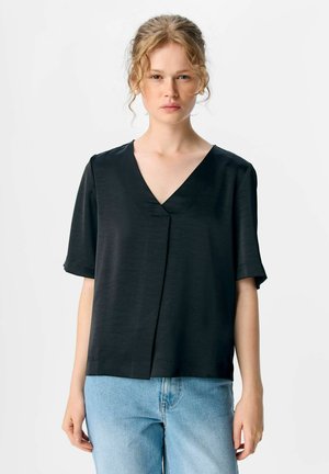 Jeune femme aux cheveux blonds bouclés portant un blouse noire à col en V, à manches courtes et un jean léger bleu, devant un fond blanc uni.