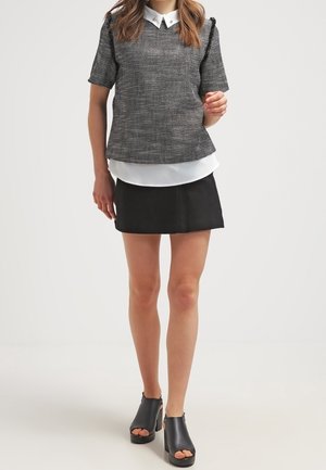 Kobieta ubrana w szary, teksturowany top z krótkim rękawem i białym kołnierzykiem, czarną mini spódniczkę oraz czarne sandały na obcasie, stojąca na białym tle.