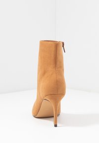 Bottine à talon haut en daim fauve avec un talon aiguille fin et une fermeture éclair à l'arrière, vue de dos sur une surface blanche.