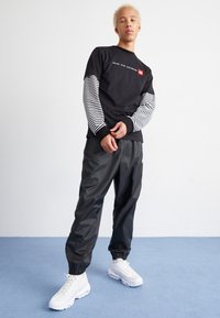 Zwarte lange mouwen shirt met gestreepte mouwen en tekst, gecombineerd met glanzende zwarte broek en witte sportschoenen, staand op een blauwe ondergrond.