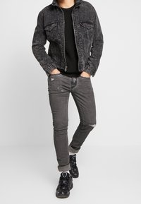 Personne portant une veste en denim noir délavé, un pull en tricot noir, un jean skinny gris usé, des chaussettes blanches et des baskets noires, se tenant avec les mains dans les poches.