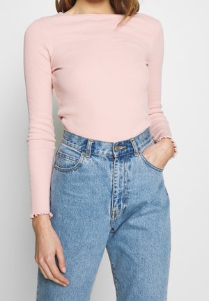 Vrouw die een lichtroze geribd shirt met lange mouwen en kroese randen bij de manchetten draagt, gecombineerd met een high-waisted lichtblauwe spijkerbroek, hand in de zak.