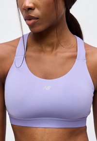 Γυναίκα με ανοιχτόμωβ αθλητικό σουτιέν New Balance, μαλλιά μαζεμένα πίσω, περικομμένη στους ώμους και στη μέση με λευκό φόντο.