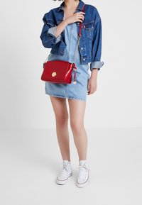 Borsa a tracolla rossa in pelle con logo dorato, dotata di catena e tracolla regolabile, texture liscia, forma rettangolare e chiusura con zip.