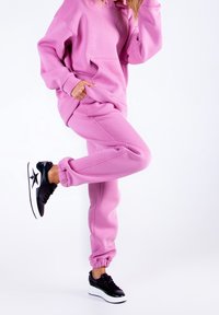 Osoba ubrana w jaskrawy różowy hoodie i pasujące dresy, unosząca jedną nogę, w czarnych sneakersach na platformie z białymi podeszwami i wzorem gwiazdy.