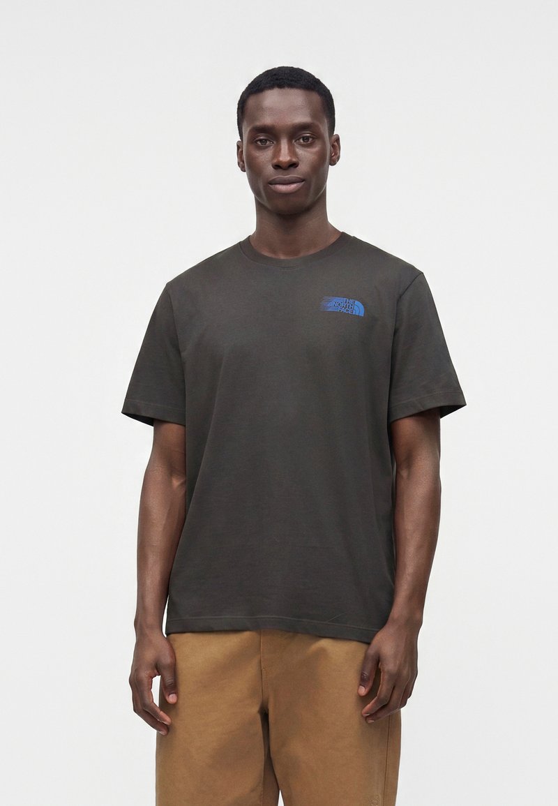 Άνδρας στέκεται φορώντας ένα σκουρογρίζο T-shirt The North Face και ανοιχτό καφέ παντελόνι μπροστά σε λευκό απλό φόντο.