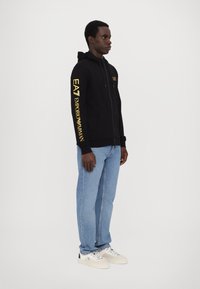 Zwarte rits hoodie met voorzakken en gouden letters op de mouwen; gecombineerd met lichtblauwe spijkerbroek en neutrale sneakers.