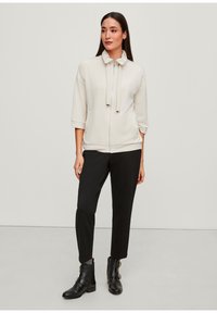 Lichtbeige zipjack met een hoge kraag, trekkers en driekwart mouwen; gecombineerd met zwarte tapered broek en zwarte enkellaarsjes.
