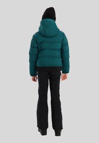 Persoon in een teal gekleurde gewatteerde jas met capuchon, zwarte broek, zwarte muts en zwarte laarzen, staand met de rug naar de camera op een lichtgrijze achtergrond.