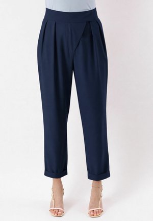 Pantaloni a vita alta color blu navy con taglio affusolato, pieghe frontali e orli risvoltati, indossati con tacchi con cinturino rosa chiaro su sfondo bianco.