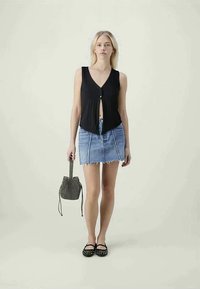Blusa preta sem mangas com decote em V e detalhe de botão, combinada com uma saia de jeans azul com bordas desfiadas e costura contratante. Sapatos planos pretos e uma bolsa estampada.