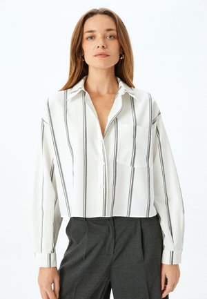 Camicia button-up tagliata in bianco con righe verticali nere, dotata di colletto, una tasca sul petto e maniche lunghe con polsini abbottonati.