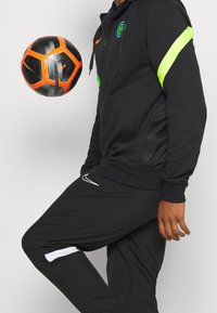 Hoodie zippé noir avec des accents vert fluo et jaune, associé à un pantalon noir avec une bande blanche ; un ballon de football noir et orange flotte à proximité.